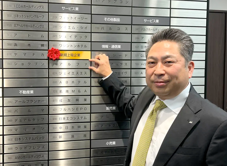 名古屋証券取引所の銘柄ボード前で新規上場した当社プレートを指差す岡本社長